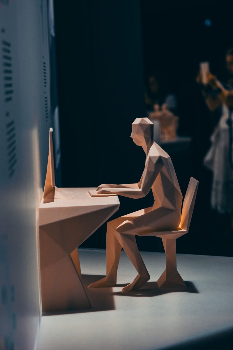 Artistic abstract sculpture of geometric figure seated at a desk, exploring technology themes.